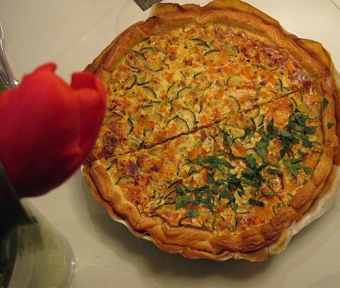 Goldbraun gebackene Quiche mit geräucherter Forelle, Zucchini und Kräutern auf einem weißen Teller serviert.