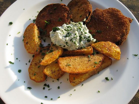 Knusprig frittierte Auberginenscheiben mit goldbraunen Kartoffelspalten und einer cremigen Knoblauch-Kräuter-Mayonnaise auf weißem Teller angerichtet, mit frischen Kräutern bestreut, warm serviert im Sonnenlicht.