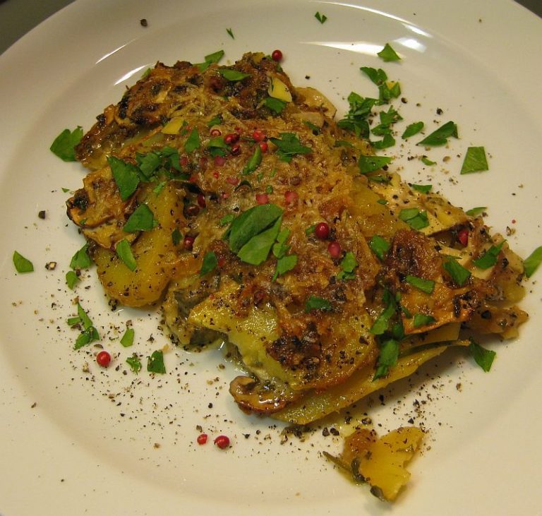 Goldbraun gebackener Kartoffel-Pilzauflauf aus Apulien mit Champignons, Parmesan, Petersilie und grobem Pfeffer auf weißem Teller serviert.