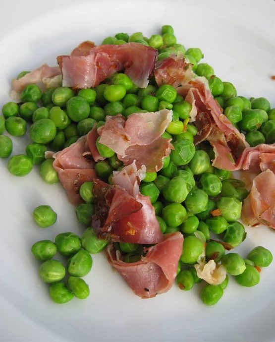 Frische grüne Erbsen mit in Streifen geschnittenem rohem Schinken auf einem weißen Teller angerichtet, leicht glänzend und schlicht präsentiert