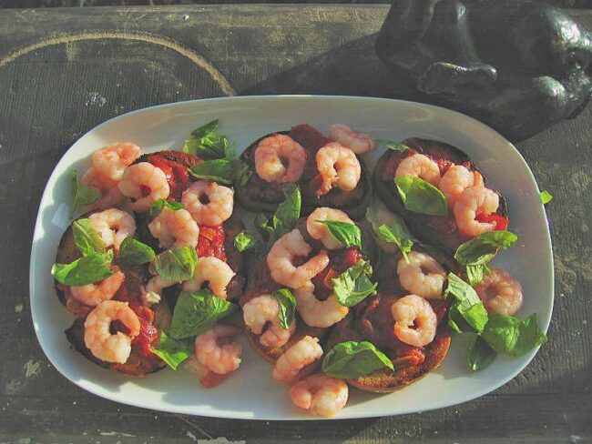 Geröstete Brotscheiben mit eingekochter Tomatensauce, Garnelen und frischem Basilikum auf weißer Platte, serviert im warmen Licht