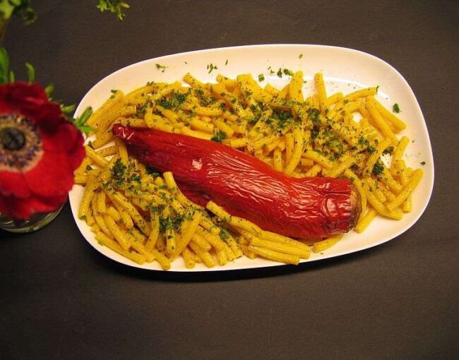 Mit Hackfleisch gefüllte rote Spitzpaprika auf gelben Kurkuma-Nudeln mit gehackten Kräutern auf eineer weißen Servierplatte.
