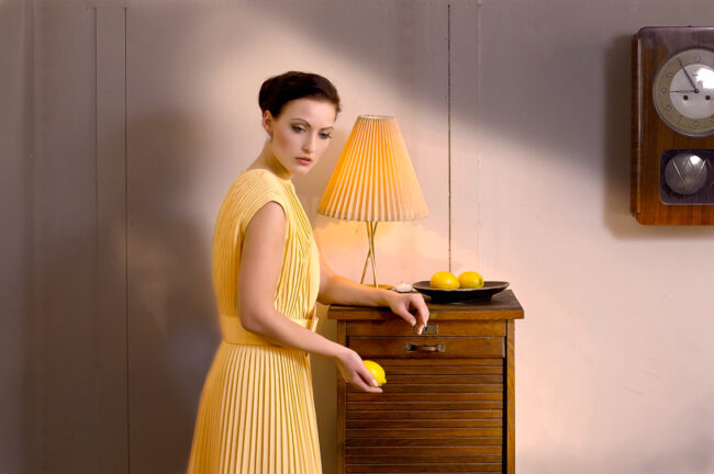 Horst Kistner almost nine am ist eine Stilles Bild im Vintage Stil. Eine Frau steht melancholisch an einem Möbel und hält eine Zitrone in der Hand.