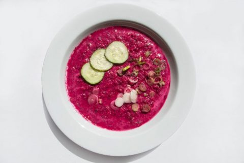 Kalte Rote-Bete-Suppe mit Gurkenscheiben und Frühlingszwiebeln in einem weißen Teller vor hellem Hintergrund.