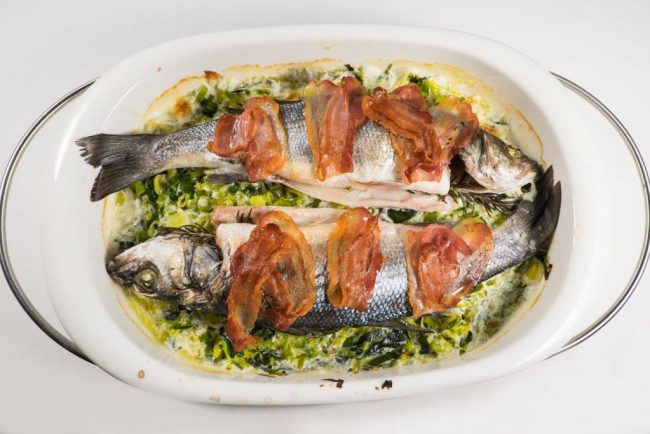 Wolfsbarsch auf Lauchgemüse mit Speck wird zusammen im Ofen gebacken. Die Speckstraeifen brät man in der Pfanne an und legt die Streifen über den Fisch