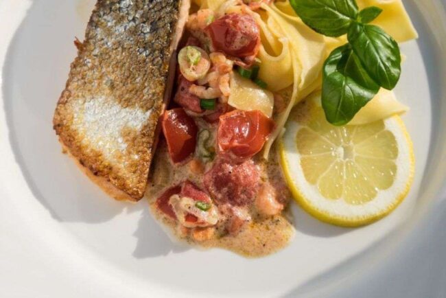 Gebratener Lachs mit knuspriger Haut, Bandnudeln und Krabbensoße mit Tomaten und Frühlingszwiebeln, dazu Basilikum und eine Zitronenscheibe.