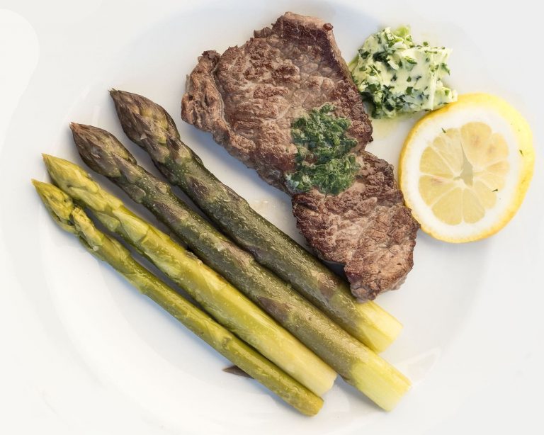 Gebratenes Bison Steak auf weißem Teller, serviert mit grünem Spargel, einer Portion Bärlauchbutter und einer halben Zitronenscheibe. Das Steak ist medium gebraten und mit geschmolzener Bärlauchbutter belegt, der Spargel liegt parallel angerichtet. Schlichte, helle Präsentation eines Frühlingsgerichts mit Bisonfleisch, Spargel und Bärlauch.