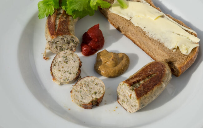 Die Hirschbratwurst mit Bärlauch im Anschnitt zeigt wie das Brät einer Bratwurst auszusehen hat. Denn das Brät soll nicht so fein gekuttert werden wie das einer Brüh- oder Bockwirst. Man erkennt deutlich die Inhaltsstoffe und die Gewürze.