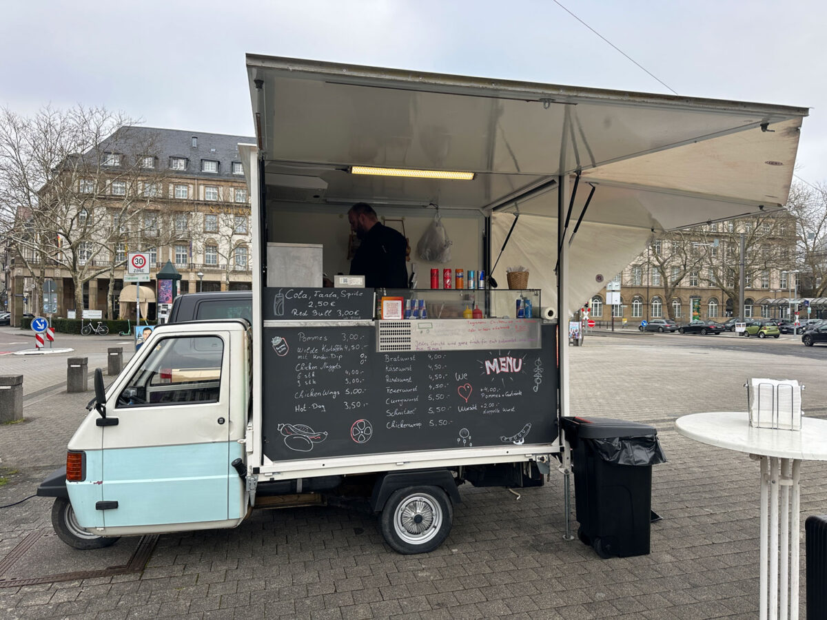 Ein skurriler Wurstwagen auf dem Karlsruher Bahnhofsvorplatz rettet diese gepflasterte Wüste zwar nicht, gibt ihr jedoch ein wenig Leben. Auf einem italienischen Piaggo einen Wurststand zu montieren ist genial.