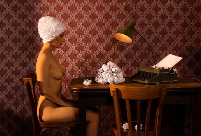 Die surreale, erotische Fotografie von Horst Kistner mit dem Titel The Paper Rose zeigt eine sitzende nackte Frau an einem Tisch mit einer Schreibmaschine und zerknülltem Papier. Sie hat eine Kappe, auf die dem Papier ähnelt.