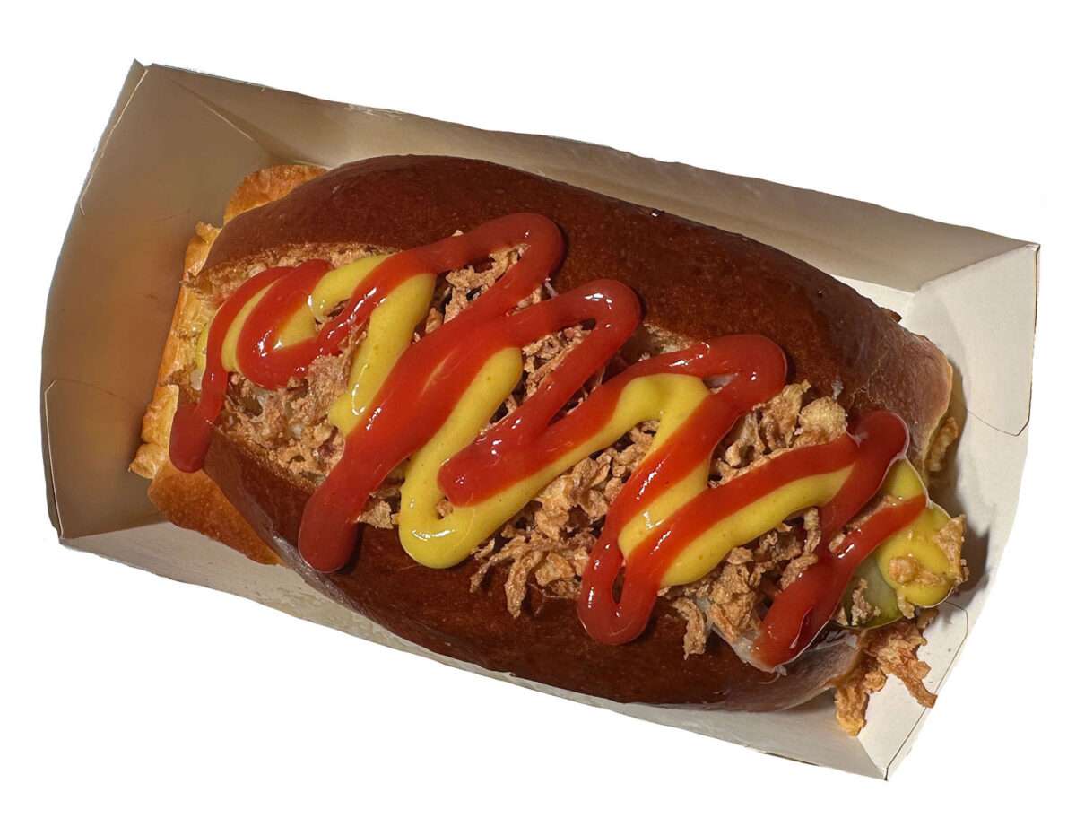 Der oder das Hamburger Hot Dog im Schlafrock im Sarg aus Pappe ist mobil, da man es wegtragen kann. Bekleckern wird man sich trotzdem, da das Teigstück für einen normalen Mund zu groß ist.