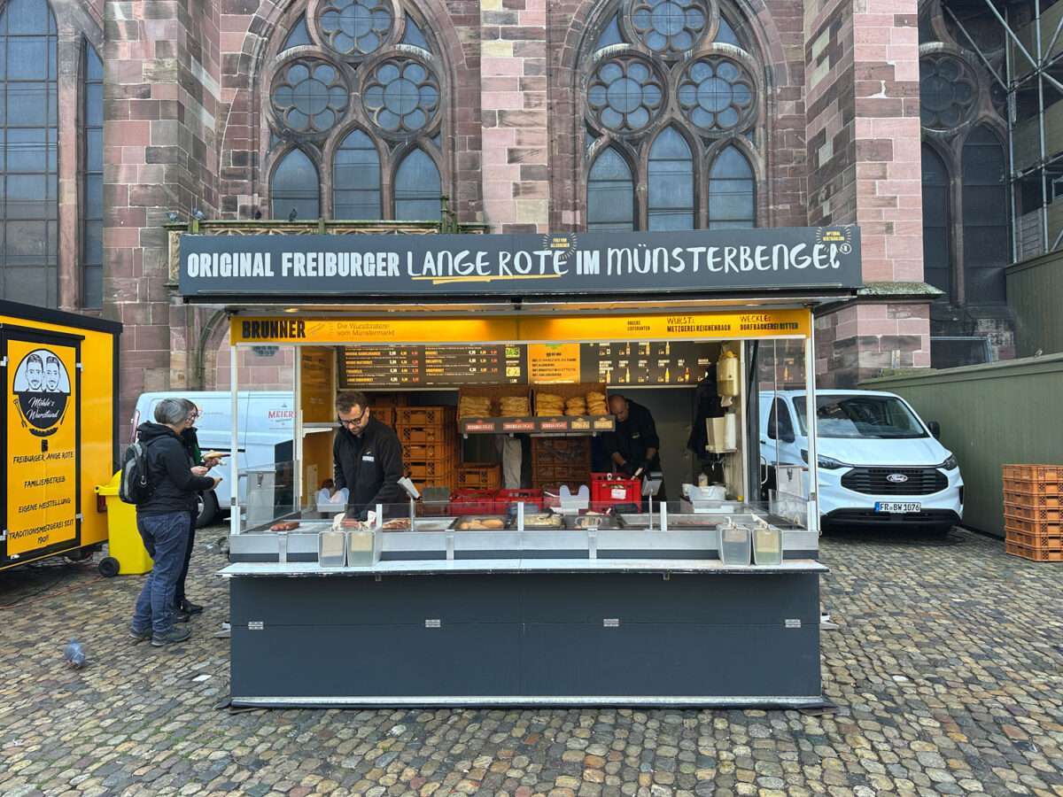Brunner-die-Wurstbräterei-vom-Münstermarkt Brunner Wurstbräterei „Original Freiburger Lange Rote im Münsterbengel“ am Münsterplatz Freiburg, Kunde an der Theke, Münster im Hintergrund.