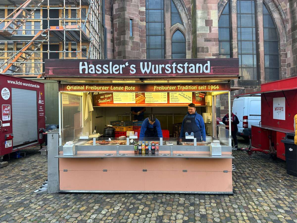 Hasslers-Wurststand-auf-dem-Münstermarkt-Freiburg Hassler’s Wurststand am Münsterplatz Freiburg vor der Münsterfassade, Schild „Original Freiburger Lange Rote“, zwei Mitarbeiter hinter der Theke und ferig gebratene Würste.