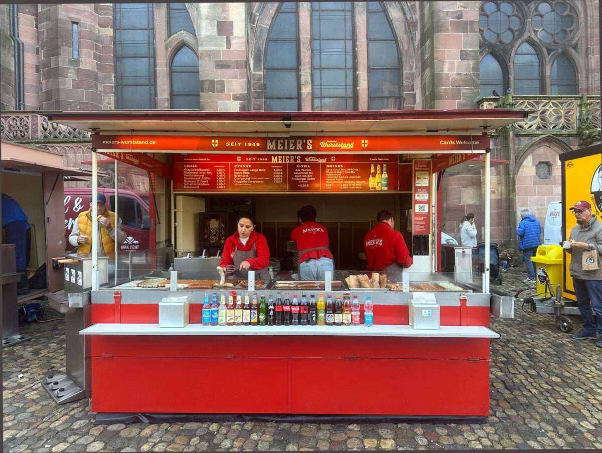Meier`s-Wurststand-auf-dem-Freiburger-Markt Meier’s Wurststand am Münsterplatz Freiburg, roter Stand mit Getränkeflaschen und Mitarbeitern in roten Jacken, Münsterfassade dahinter