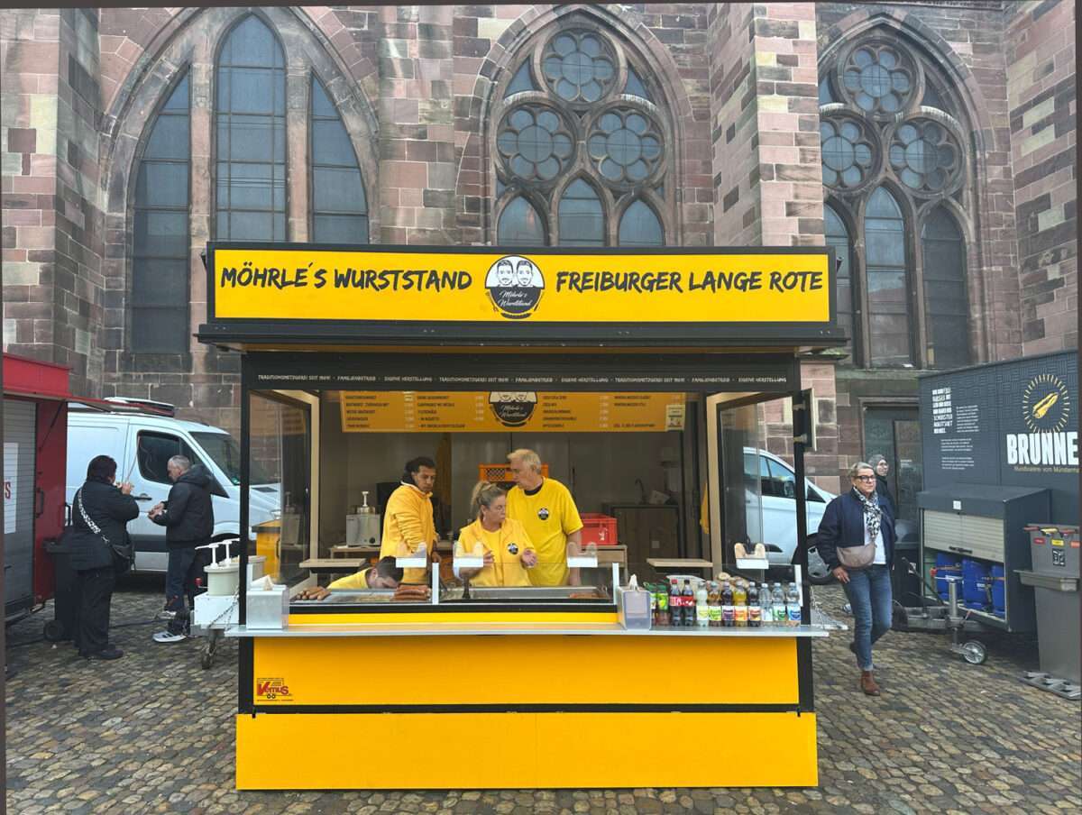 Möhrle`s-Wuststand-auf-dem-Freiburger-Markt Möhrle’s Wurststand „Freiburger Lange Rote“ am Münsterplatz Freiburg, gelber Stand mit Team in gelben Jacken vor dem Münster.