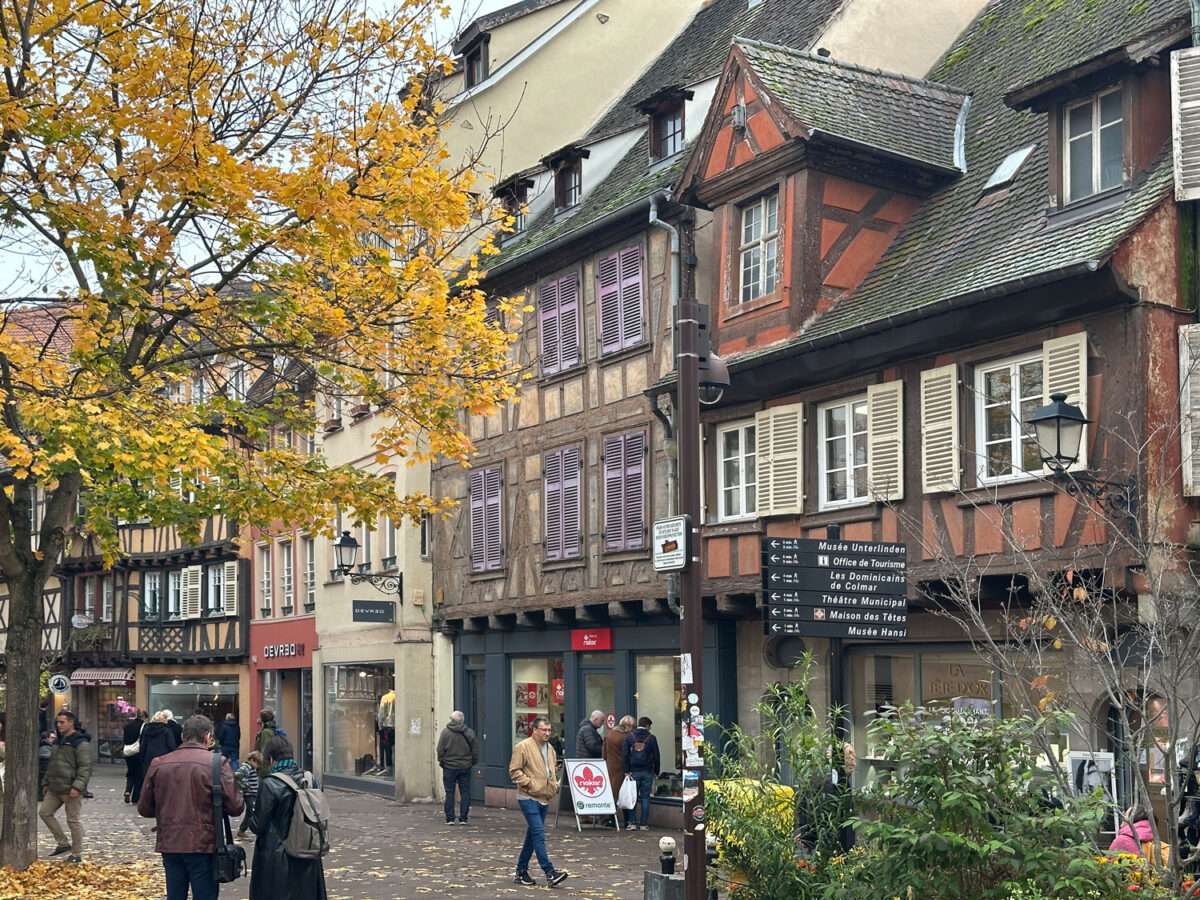 Platz in der Altstadt von Colmar mit Fachwerkhäusern, herbstlichem Baum, Passanten, Geschäften im Erdgeschoss und Wegweisern zu Museen und Sehenswürdigkeiten nahe der Maison des Têtes.