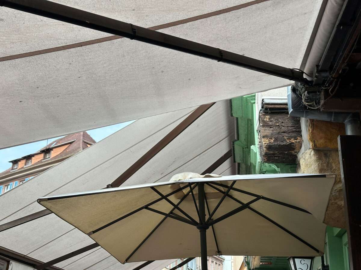 Blick nach oben aus einem Bistro in der Altstadt von Colmar mit überlappenden hellen Markisen, einem Sonnenschirm, Metallstreben, Hausfassaden und schmalem Himmelausschnitt.