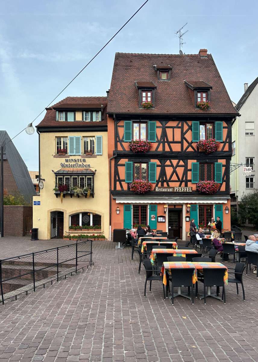 Außengastronomie auf einem gepflasterten Platz in der Altstadt von Colmar: Tische vor dem Fachwerkhaus des Restaurants Pfeffel und der Winstub Unterlinden, Gäste sitzen im Freien, historische Fassaden mit grünen Fensterläden und Blumenschmuck, ruhige Stadtszene am frühen Abend.