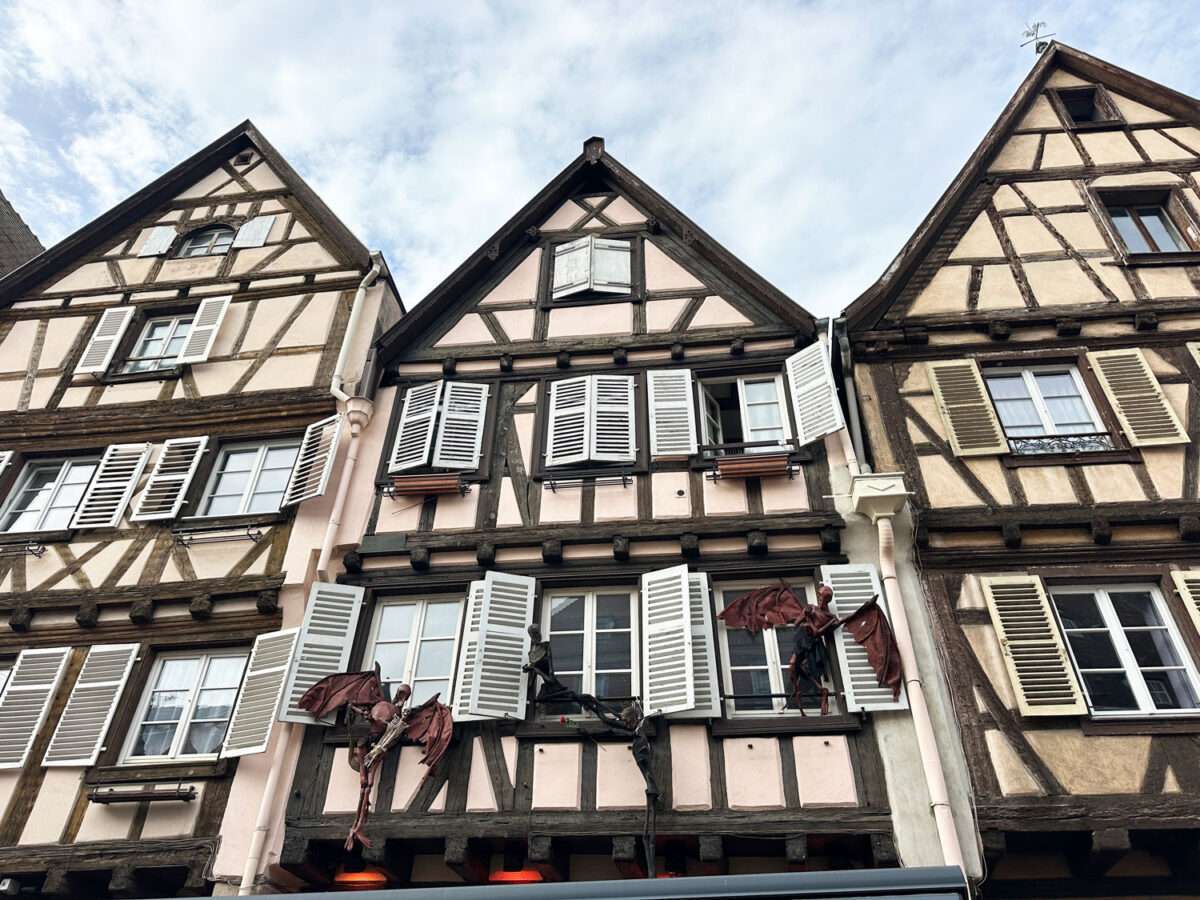 Drei eng stehende Fachwerkhäuser in der Altstadt von Colmar mit steilen Giebeln, hellen Putzfeldern und geöffneten Holzläden; unter den Fenstern sind dunkle, skelettartige Figuren mit ausgebreiteten Flügeln als auffällige Fassadendekoration angebracht.