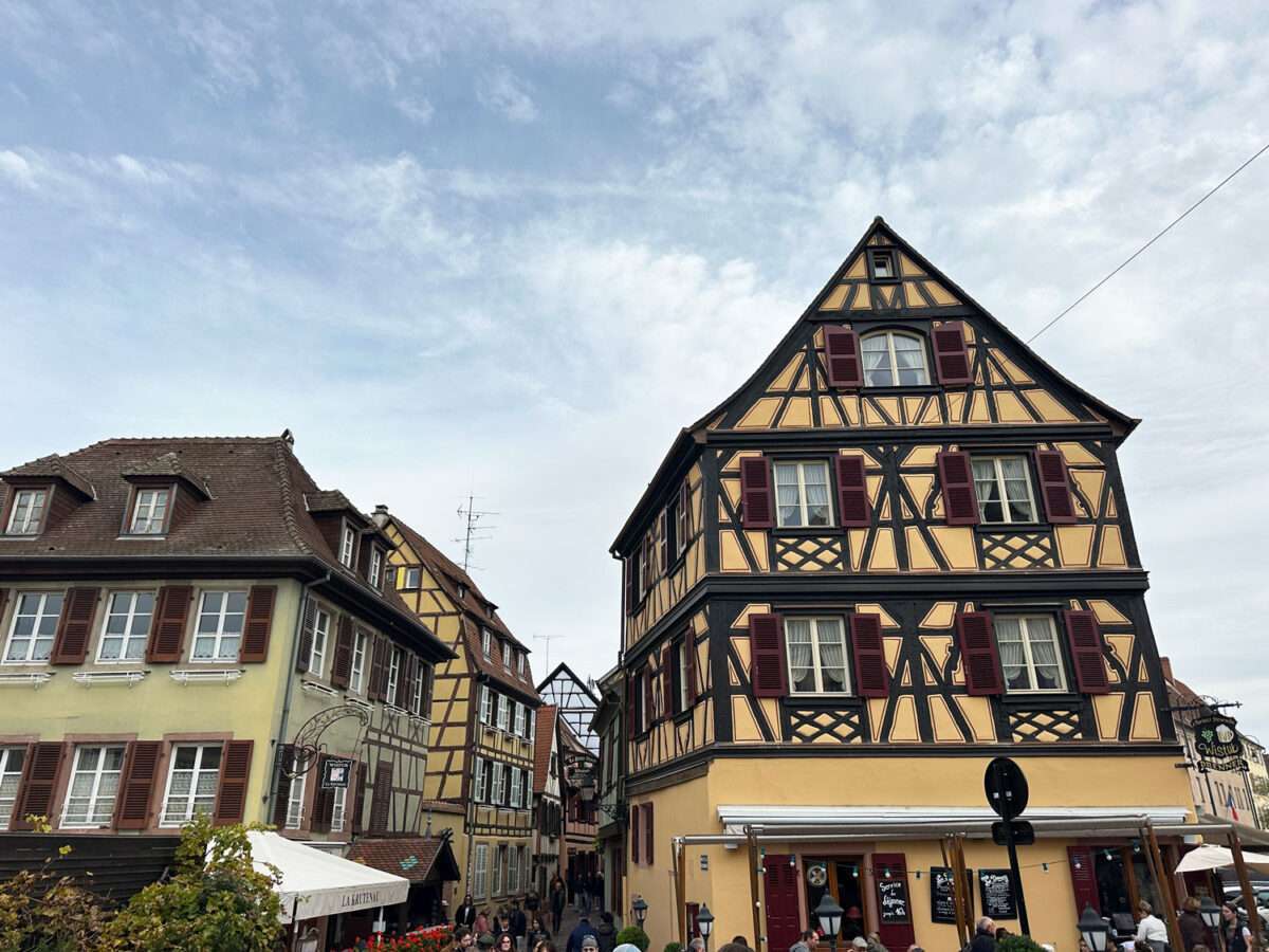 Altstadt von Colmar mit farbigem Fachwerk-Eckhaus in der Rue des Têtes, umliegenden historischen Häusern, Fensterläden, Gastronomie im Erdgeschoss und belebter Straßenszene.