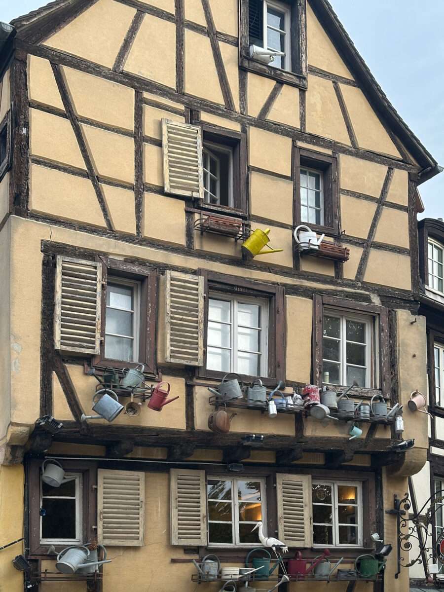 Fachwerkhaus in der Altstadt von Colmar mit ungewöhnlichem Fassadenschmuck: Zahlreiche Gießkannen aus Metall und Kunststoff sind auf Ablagen unter den Fenstern arrangiert, helle Putzflächen und dunkles Holztragwerk prägen die Fassade, typisch elsässische Architektur mit dekorativem Zierrat.