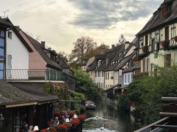 Kanal der Petite Venise in Colmar mit dicht stehenden Fachwerk- und Wohnhäusern, Restauranttischen am Wasser, Schwänen auf dem Kanal und ruhiger Altstadtatmosphäre.