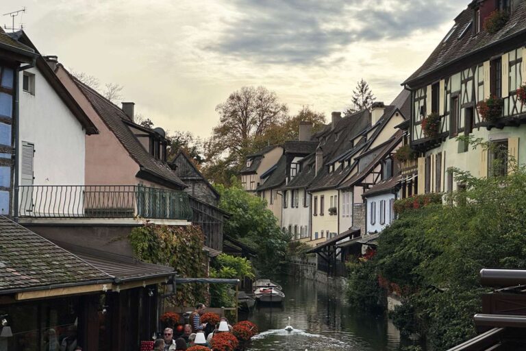 Kanal der Petite Venise in Colmar mit dicht stehenden Fachwerk- und Wohnhäusern, Restauranttischen am Wasser, Schwänen auf dem Kanal und ruhiger Altstadtatmosphäre.