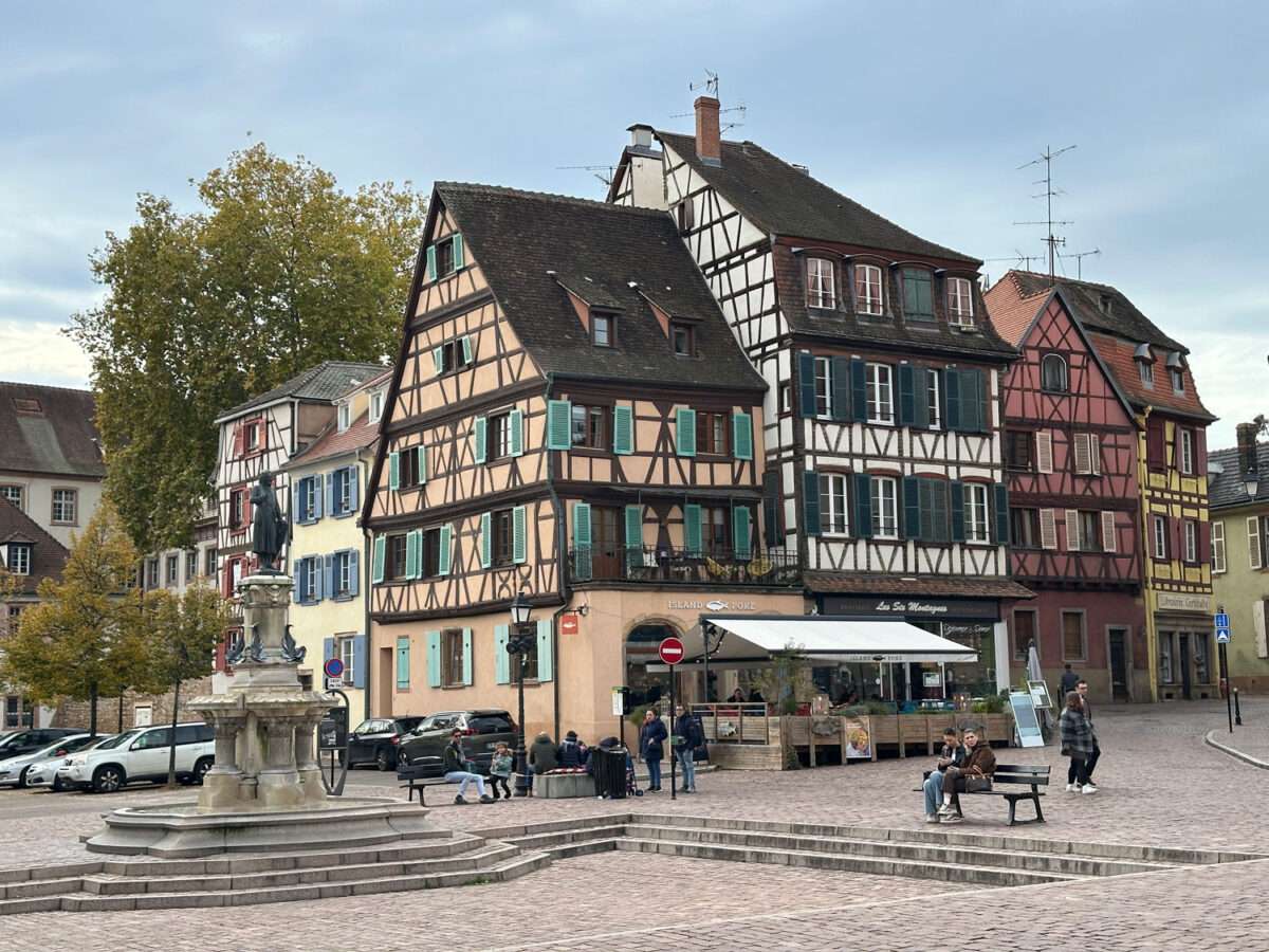 Place de l’Ancienne Douane in Colmar mit der Fontaine Schwendi im Vordergrund, dahinter farbige Fachwerkhäuser mit Fensterläden und Restaurantterrasse; auf der Markise steht „Les Six Montagnes“, auf der Fassade „Island Poké“.