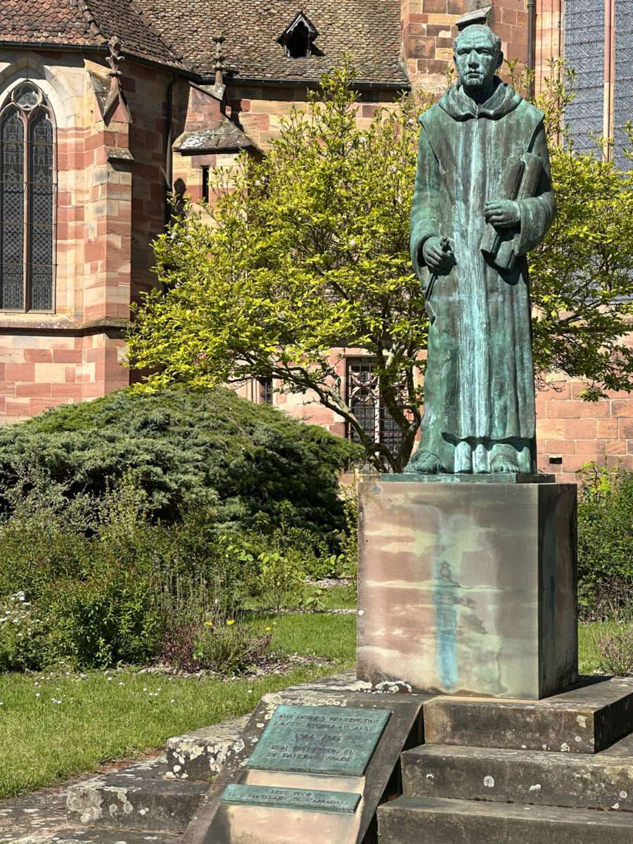 Bronzestatue eines Benediktinermönchs vor der Kirche St. Peter und Paul in Wissembourg auf Sockel mit Garten und Kirchenfassade im Hintergrund