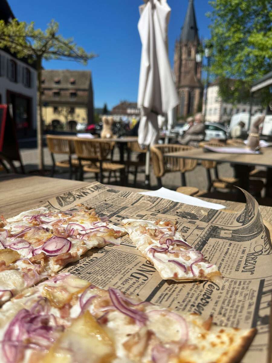 Flammkuchen auf Papier serviert in einem Bistro in Wissembourg im Elsass, mit Zwiebeln und Speck, im Hintergrund Terrasse mit Kirche