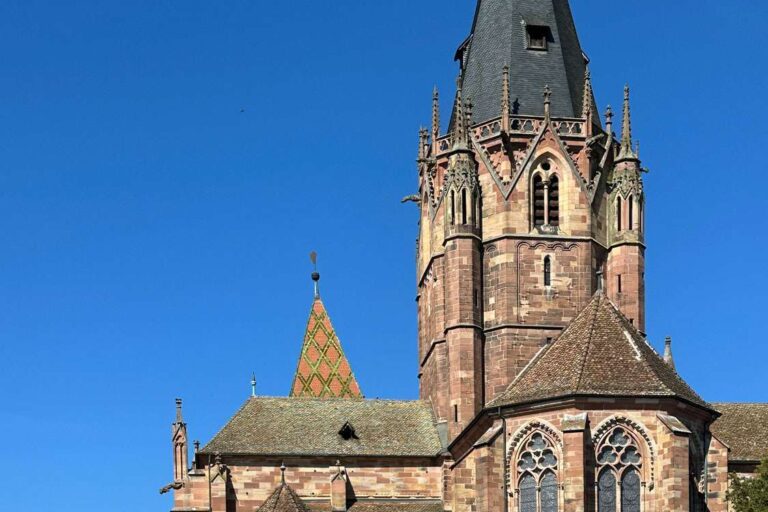 Kirche St. Peter und Paul in Wissembourg mit hohem gotischem Turm aus rotem Sandstein über den Dächern der Stadt unter blauem Himmel