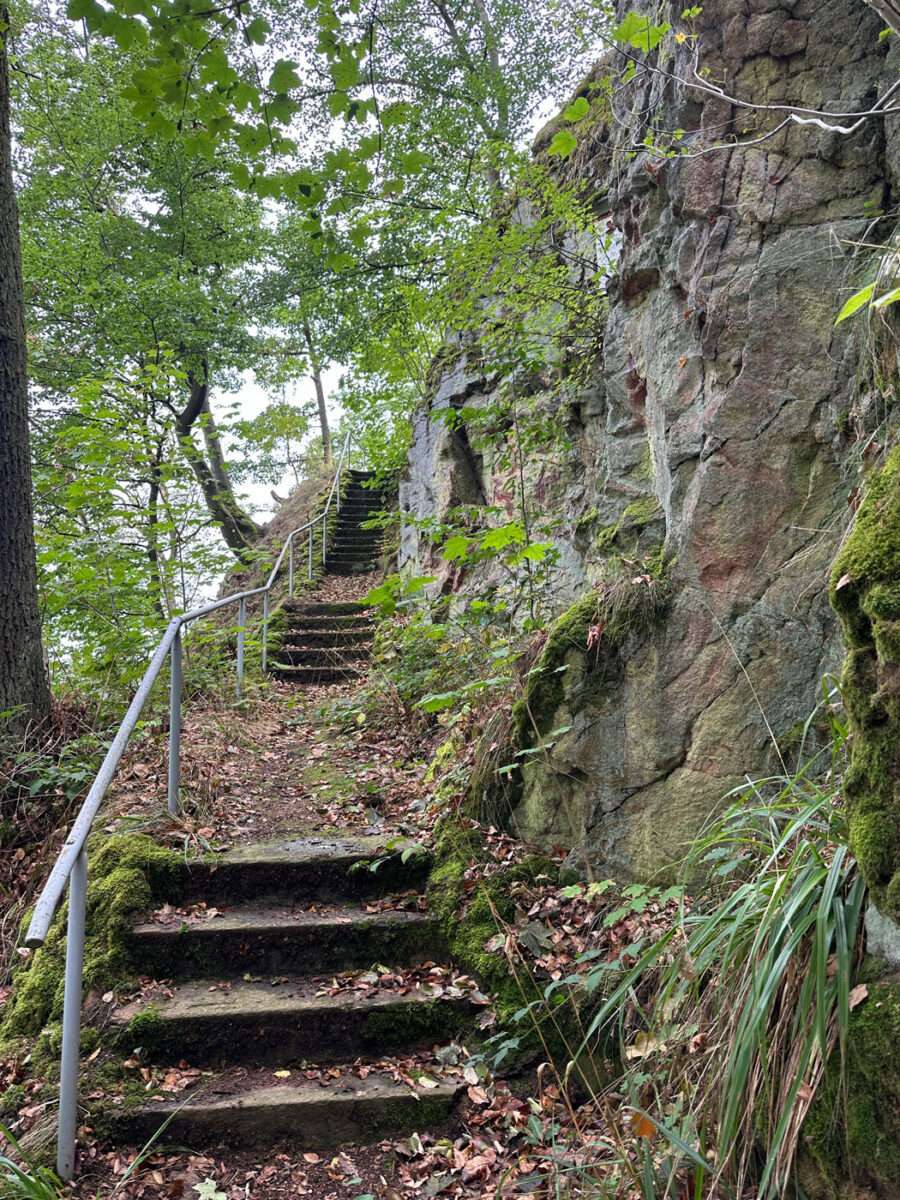 Moosbewachsene Steintreppe mit Metallgeländer entlang einer steilen Porphyrfelswand im Wald