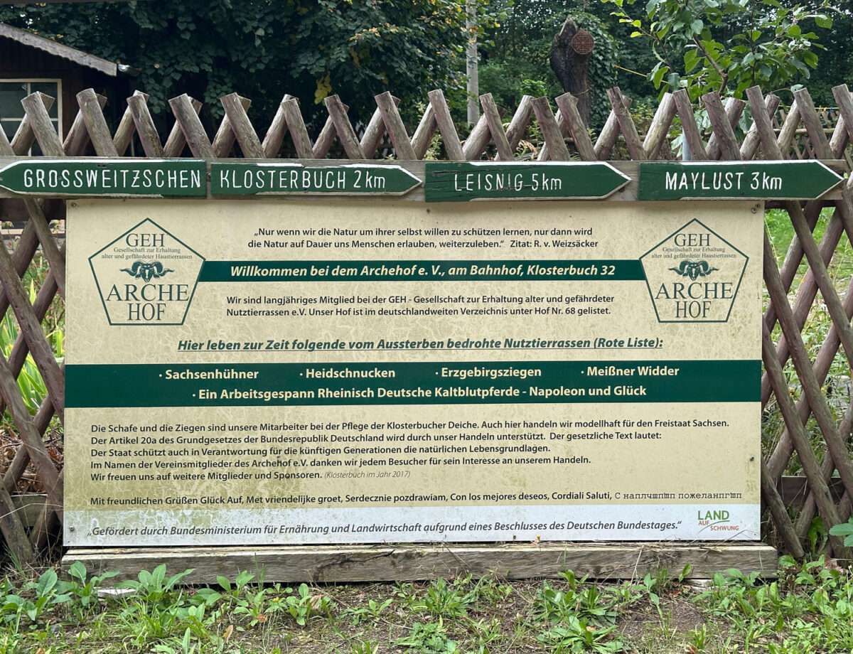 Hölzerne Wegweiser nach Großweitzschen, Klosterbuch, Leisnig und zur Maylust über einer Infotafel des Archehofs