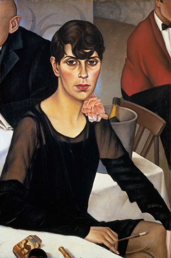 christian-schad-sonja-neue-frau-zigarettenspitze-1928 Kurzhaarige Frau im dunklen Kleid mit Zigarettenspitze am Tisch. Eine Camel Zigarettenschachtel liegt auf dem Tisch und Zwei Männer sitzen im Hintergrund.