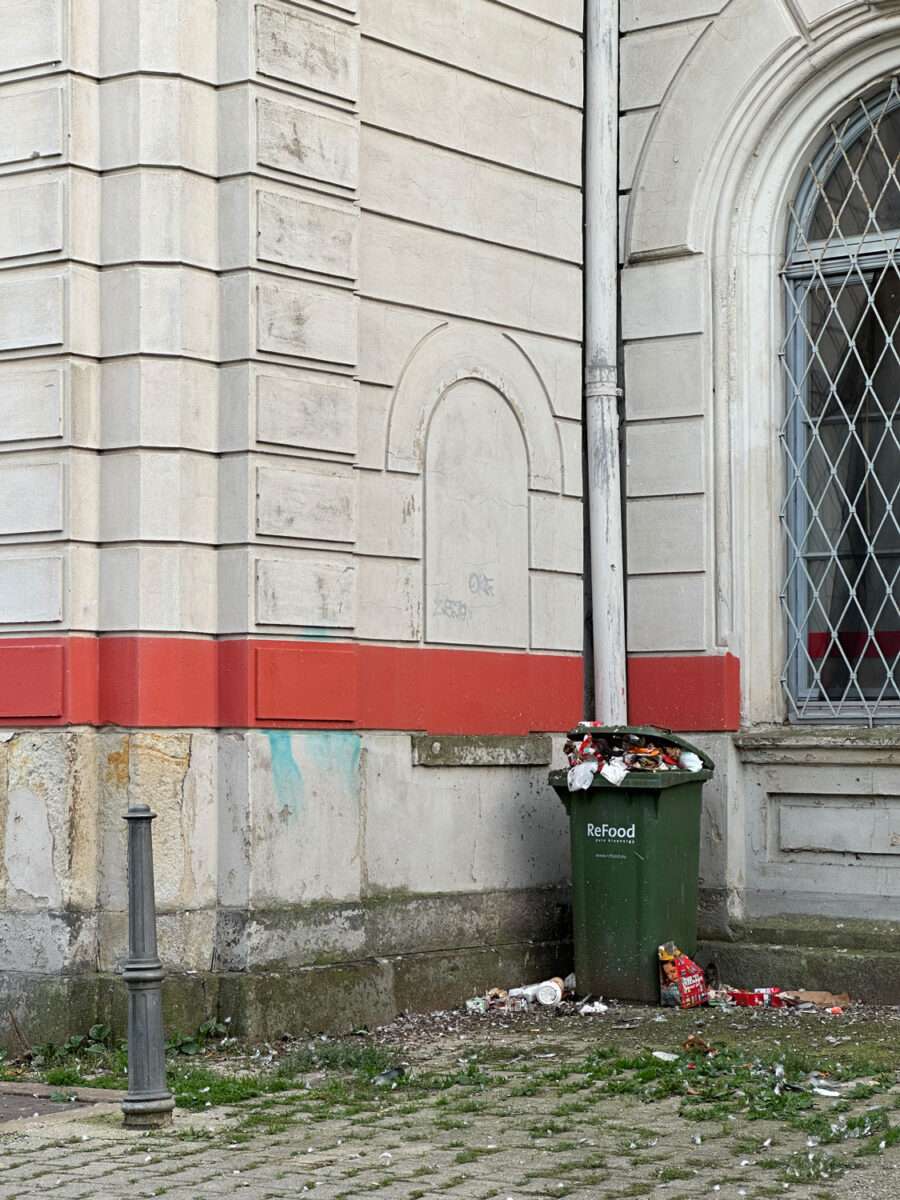 Überfüllter grüner Abfallbehälter und verstreuter Müll vor der hellen Fassade des Döbelner Hauptbahnhofs.