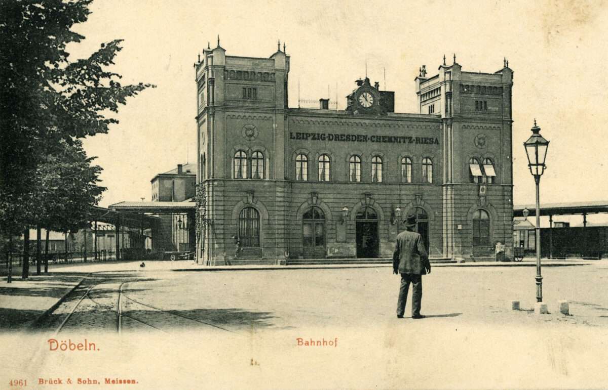 Historische Ansichtskarte des Döbelner Hauptbahnhofs mit repräsentativem Empfangsgebäude und einer Person auf dem Vorplatz.